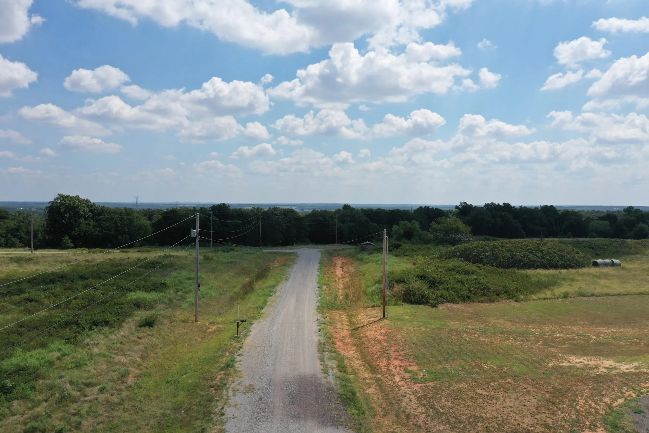 Gravel country road cutting through green Oklahoma acreage with utility poles and low brush under a bright blue sky with scattered clouds, illustrating rural land for sale and in-house financing for Shawnee acreage ownership
