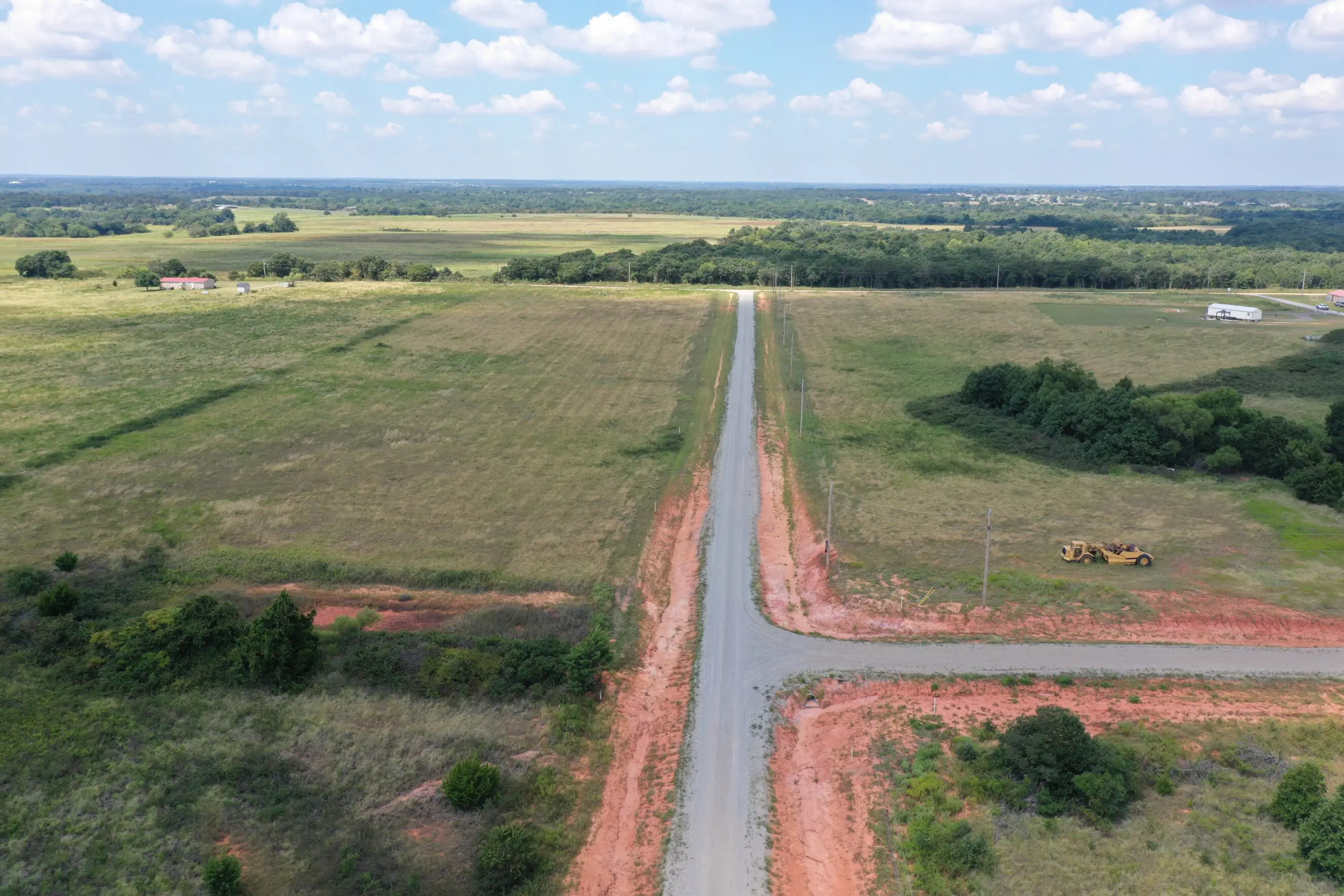 Aerial view of rural Oklahoma acreage with a straight gravel road and red clay shoulders forming a crossroad through open pasture, scattered trees, utility poles and a parked grader, suggesting ready-to-develop land and in-house financing opportunities for manufactured homes or new construction near Shawnee, OK